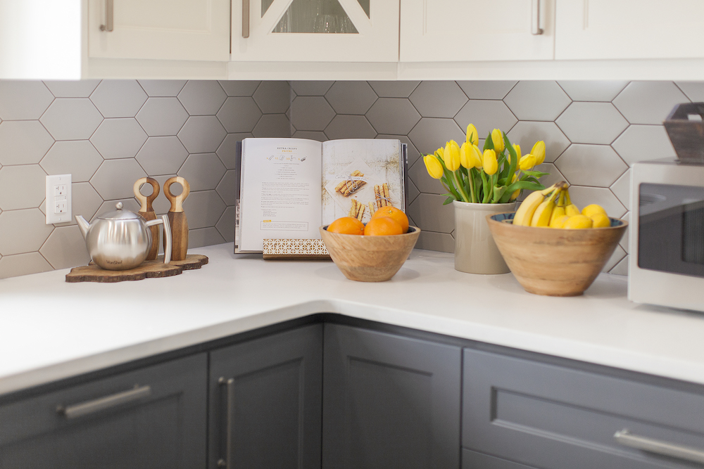 Modern kitchen with grey blue lower cabinets, white hexagon tile backsplash and a white countertop covers in wooden bowls filled with fruit and an opened cookbook