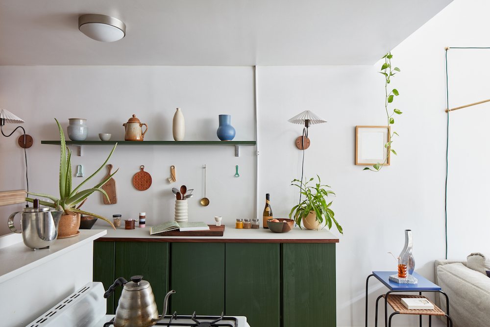 Interior of chic minimalist kitchen with a stove in the foreground and hunter green lower cabinets covered in pottery and plants in the background
