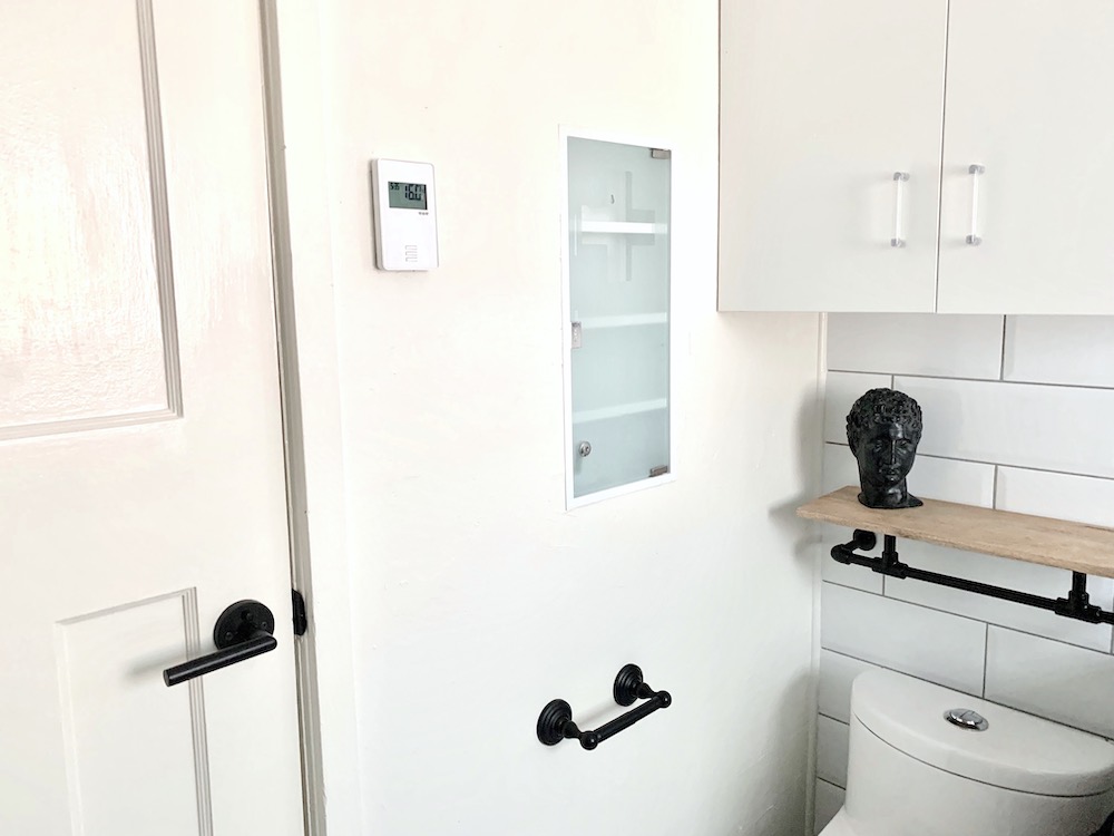 Beautiful modern bathroom with recessed medicine cabinet, white walls, white toilet, black toilet roll holder, industrial floating shelf, white door with black hands, and a large white wall cabinet