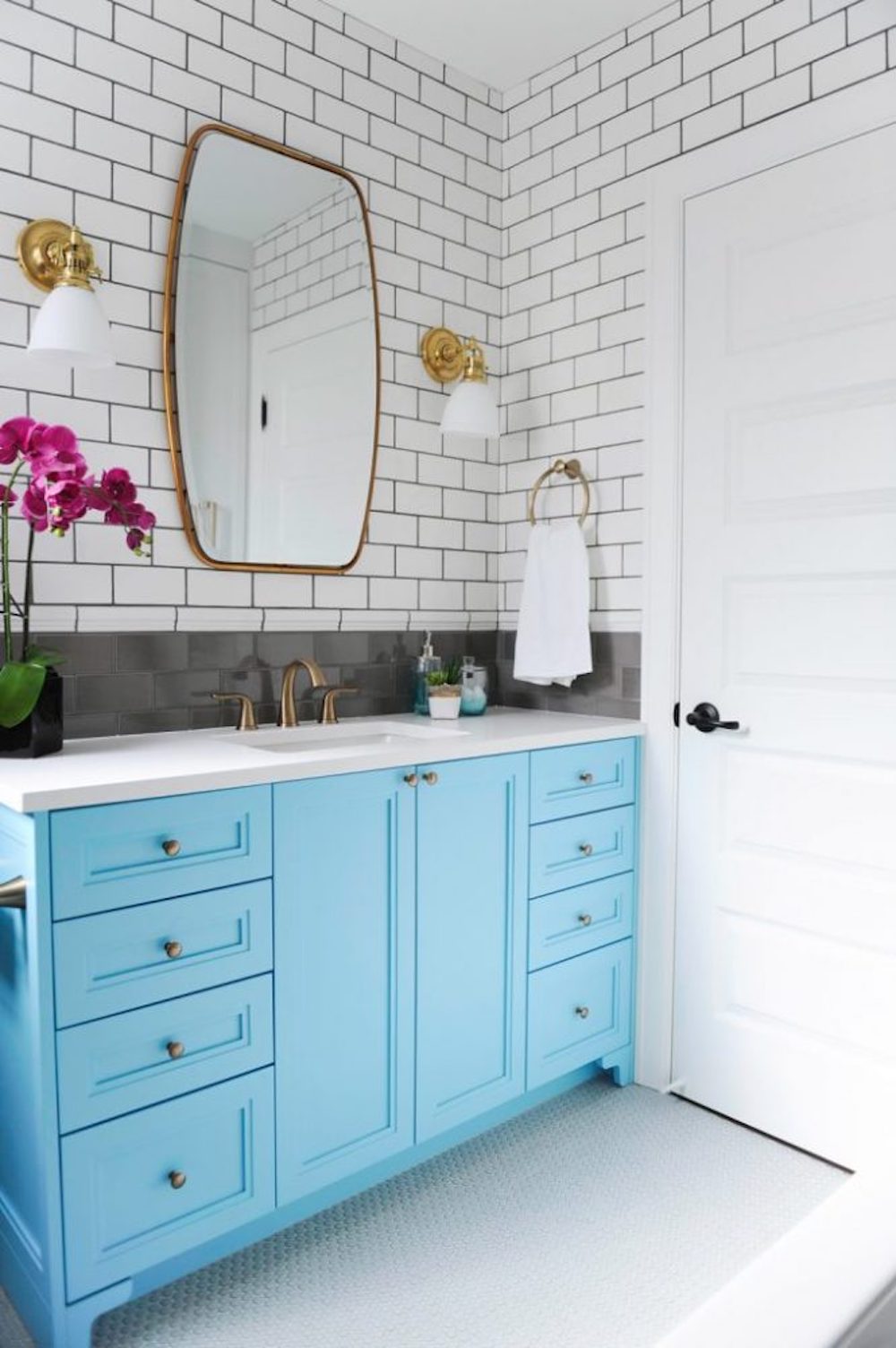 Fun and modern bathroom with white subway tiles on the wall, two brass scones, and a brass framed mirror hanging on the wall above an eggshell blue bathroom vanity