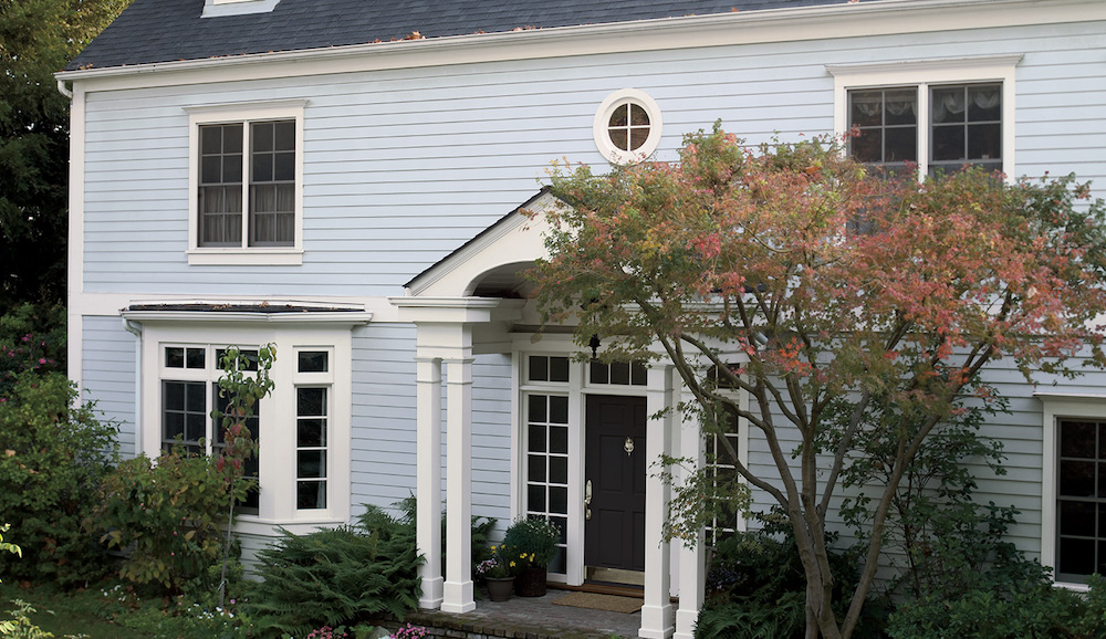 Charming blue colonial house with mature landscaping and trees, a black front door painted in BEHR Black BLACK, trim in BEHR Ultra Pure White® PPU18-06, and the main wooden siding in BEHR Ice Drop M530-1