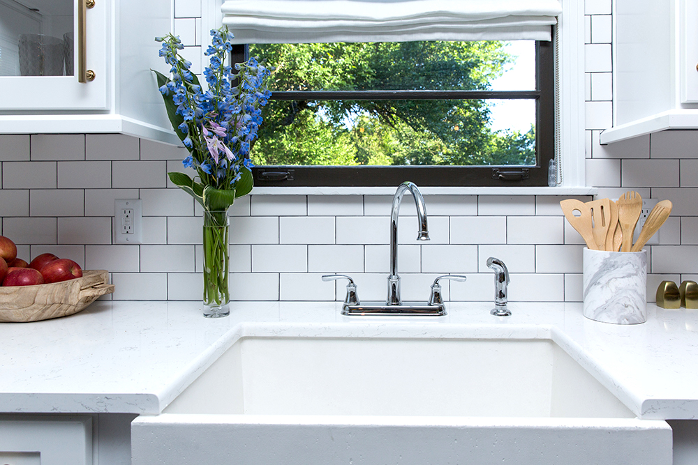 Clean white kitchen sink area with white apron sink, silver hardware, white countertops with a bowl of apples, a vase of blue flowers and a stone vessel filled with wooden utensils, a black framed window looking out onto a tree, and a white subway tile backsplash with grey grout