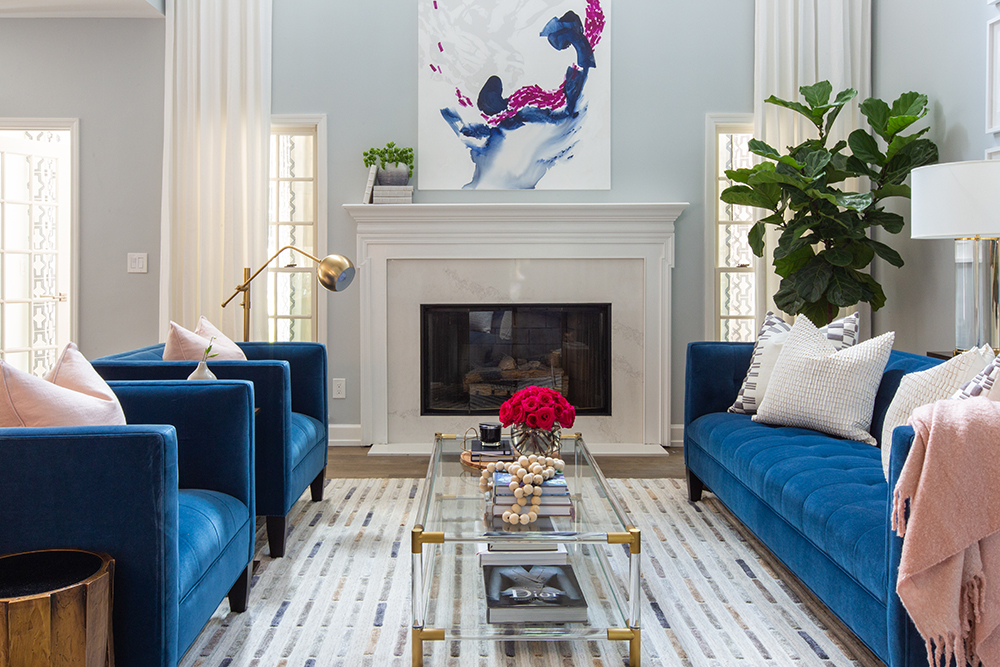Chic living room with two blue velvet armchairs, a tufted blue velvet couch, a gold floor lamp, and a gas fireplace with a white stone surround and a bold blue, fuchsia and white painting above it