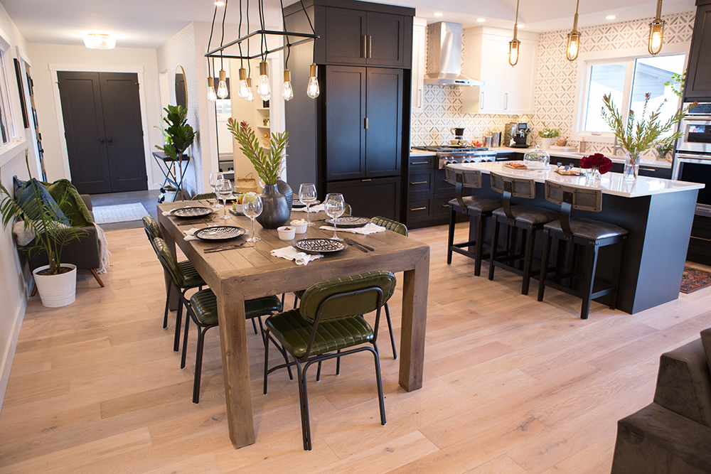 Chic modern dining room featured on The Property Brothers on HGTV with a large wooden table, six dark green vintage inspired dining chairs, a long industrial overhead chandelier, next to a large kitchen with black cabinets and a long eat-in island