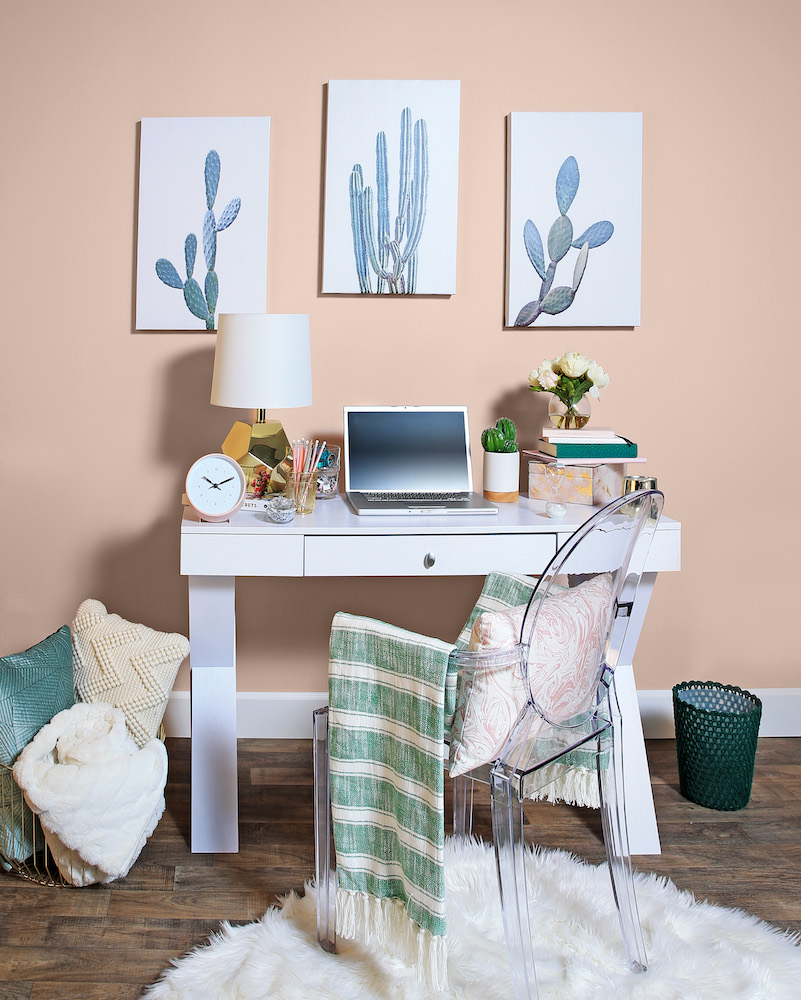 Cute office set up in a bedroom with a small white desk, acrylic chair, and three cactus prints hanging on a wall painted in BEHR’s Sand Dance S190-2 interior paint colour