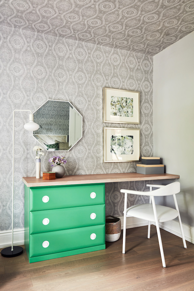 A bedroom with a funky vanity desk made from a green dresser and a long butcher block top, a modern white chair, an octagon shaped mirror, and two framed paintings