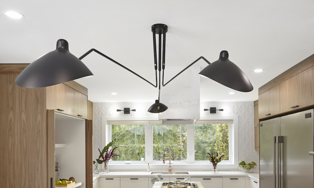 A futuristic black three shade light fixture sits in a modern kitchen with bleached oak cabinets and white countertops