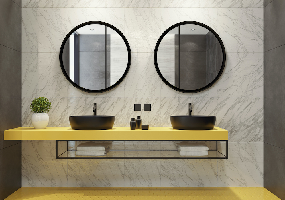 Very modern bathroom with yellow floors, marble tile on the wall, a long yellow floating vanity with two black vessel sinks and two large round mirrors on the wall about them