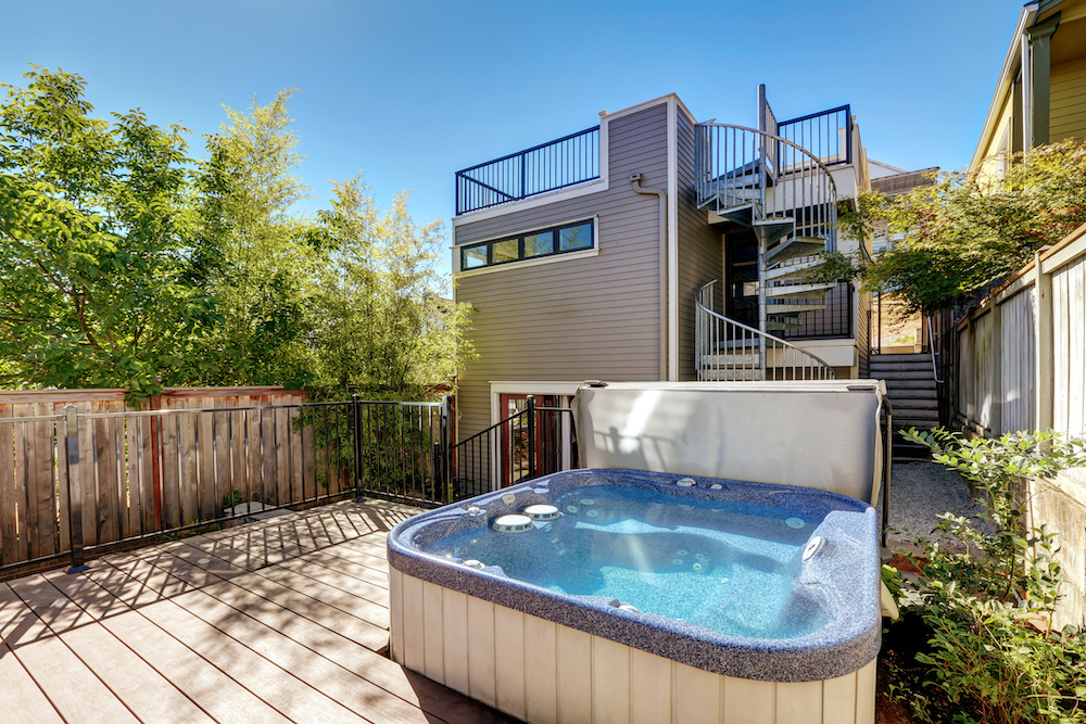 An above ground hot tub on a wooden deck below a modern grey house with a spiral staircase
