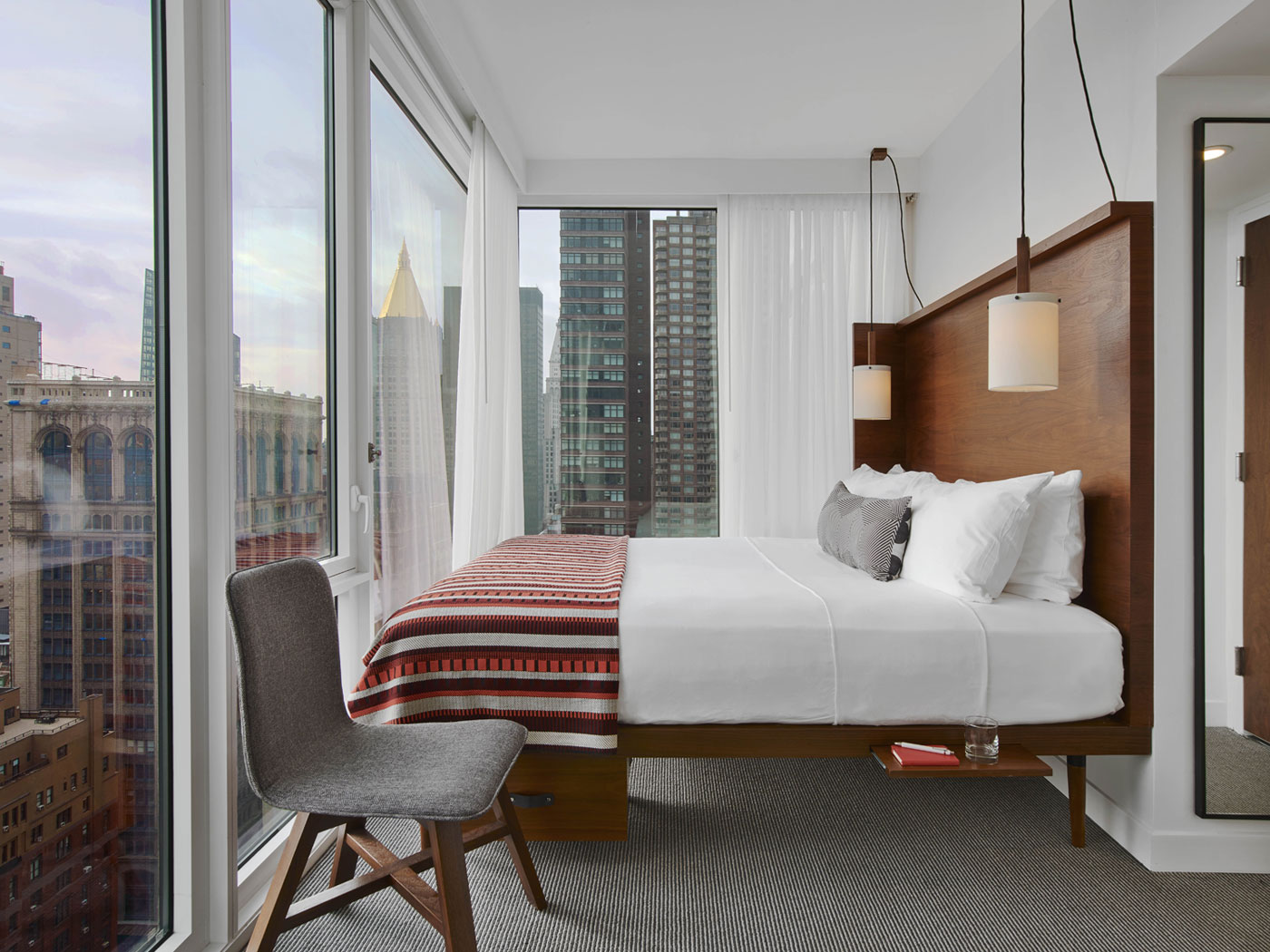 wood framed bed in small glass walled NYC hotel room with grey felt chair.