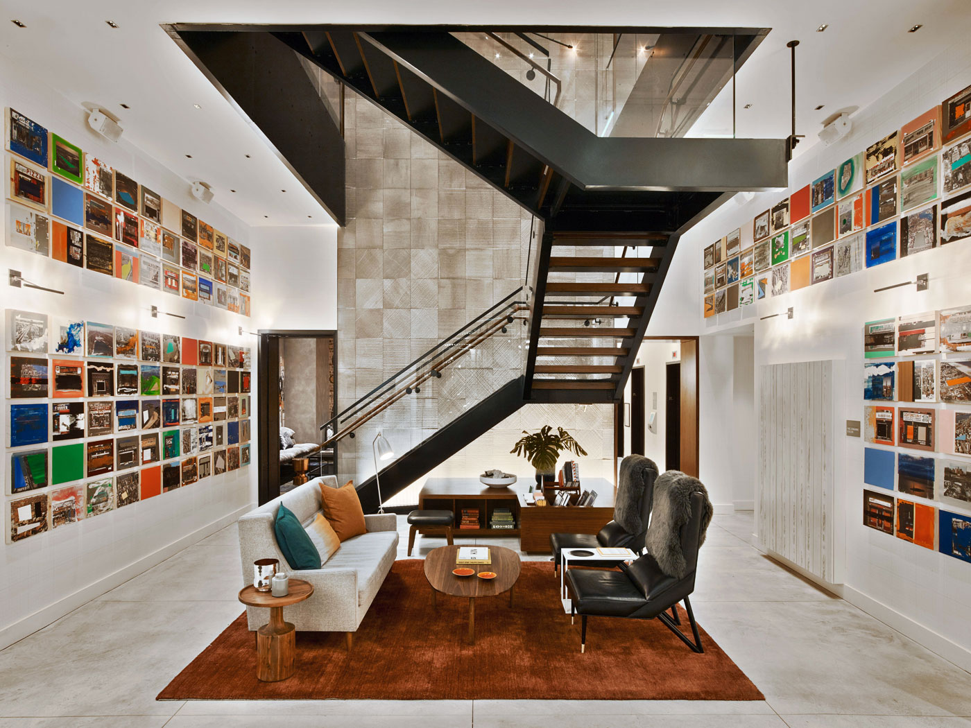 lobby with suspended staircase, rust-brown area rug with sofa and two black leather chairs and colourful artwork of the same size on both walls