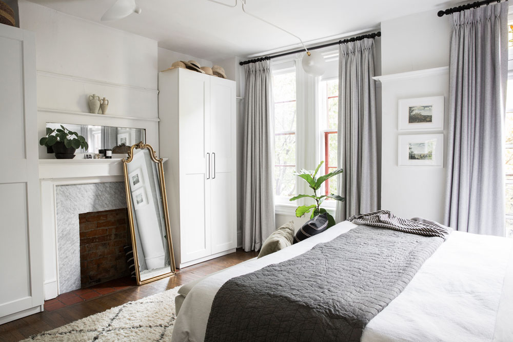 white bedroom with two windows with grey drapery and mirror on floor in front of fireplace