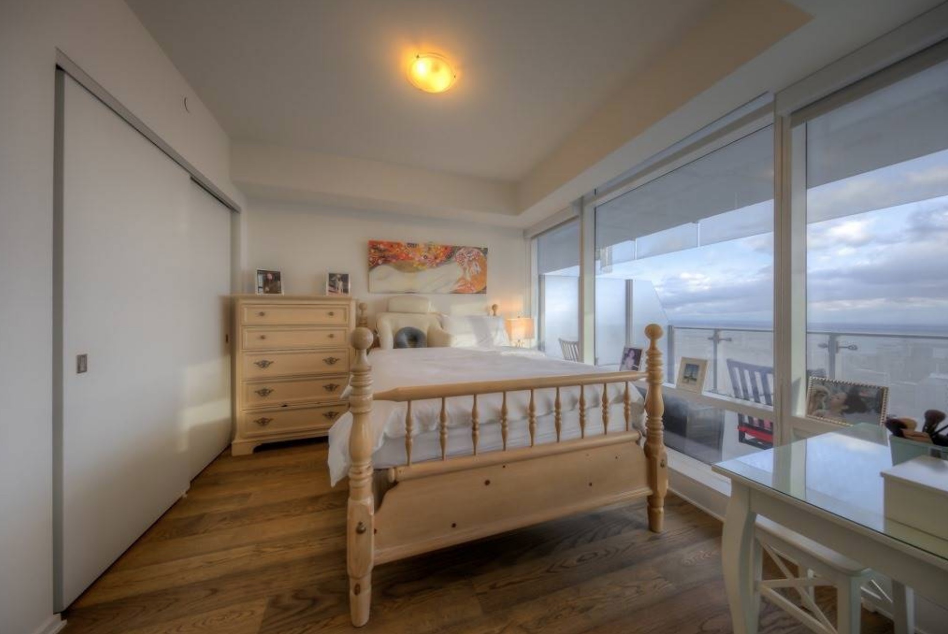 A narrow condo bedroom with hardwood flooring, wood furniture and floor-to-ceiling window overlooking the city