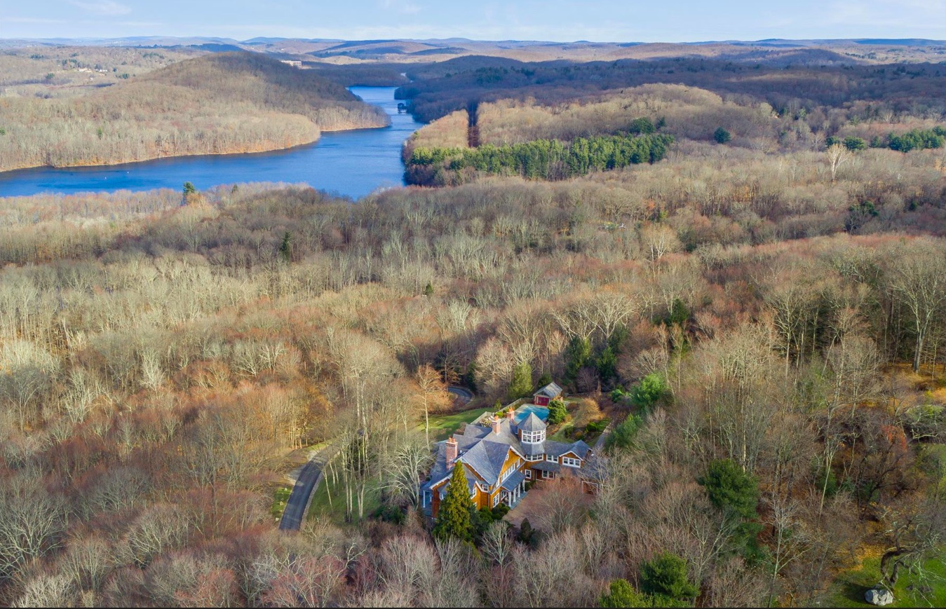 An aerial shot of the house surrounded by wooded area and body of water in the distance