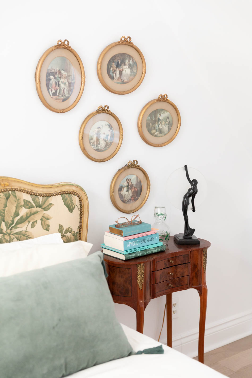 A bedside table with vintage paintings in gold frames and a stack of books