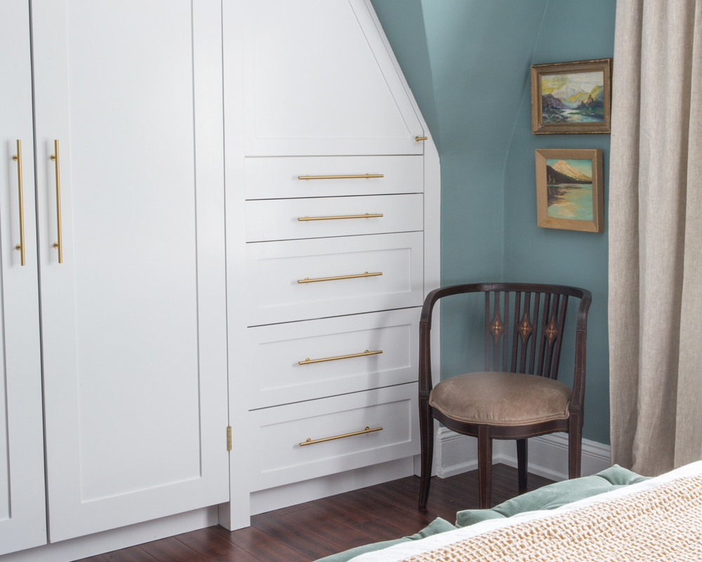 built in white cupboard and drawers with brass handles in blue green bedroom and old chair in corner