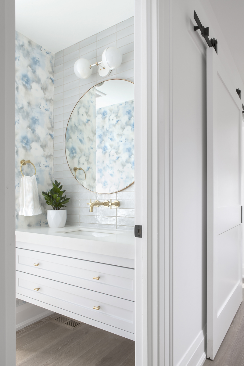 A powder room with a wallpapered accent wall and brass faucet and cupboard handles