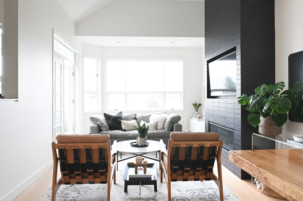 living space, back of two tan and black chairs, black fireplace, grey sofa by window