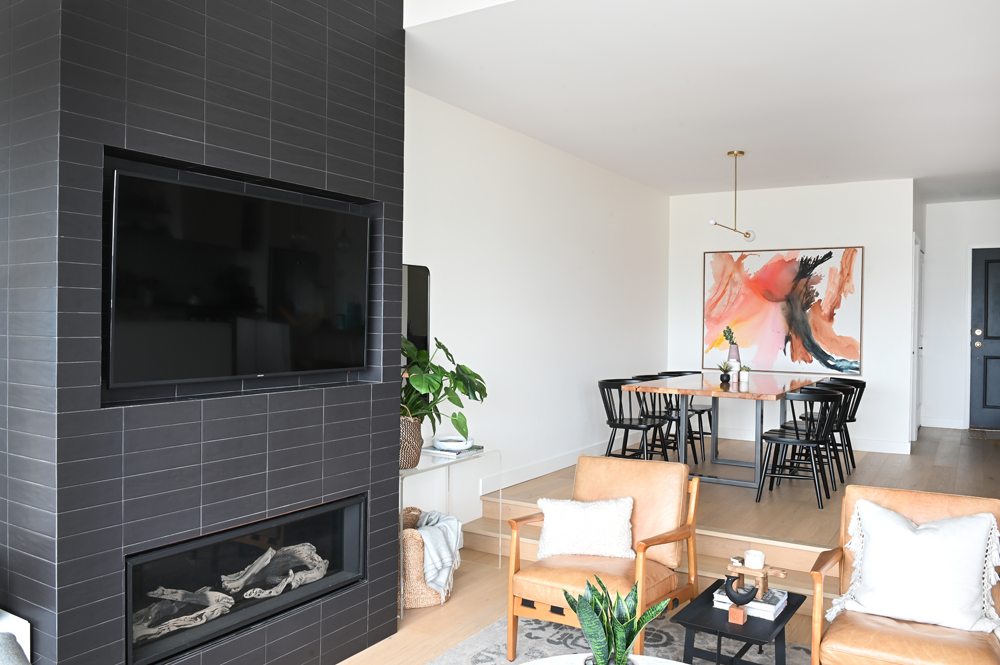 grey-black fireplace and tv living area with two tan chairs and step up to dining area with pink and black abstract art