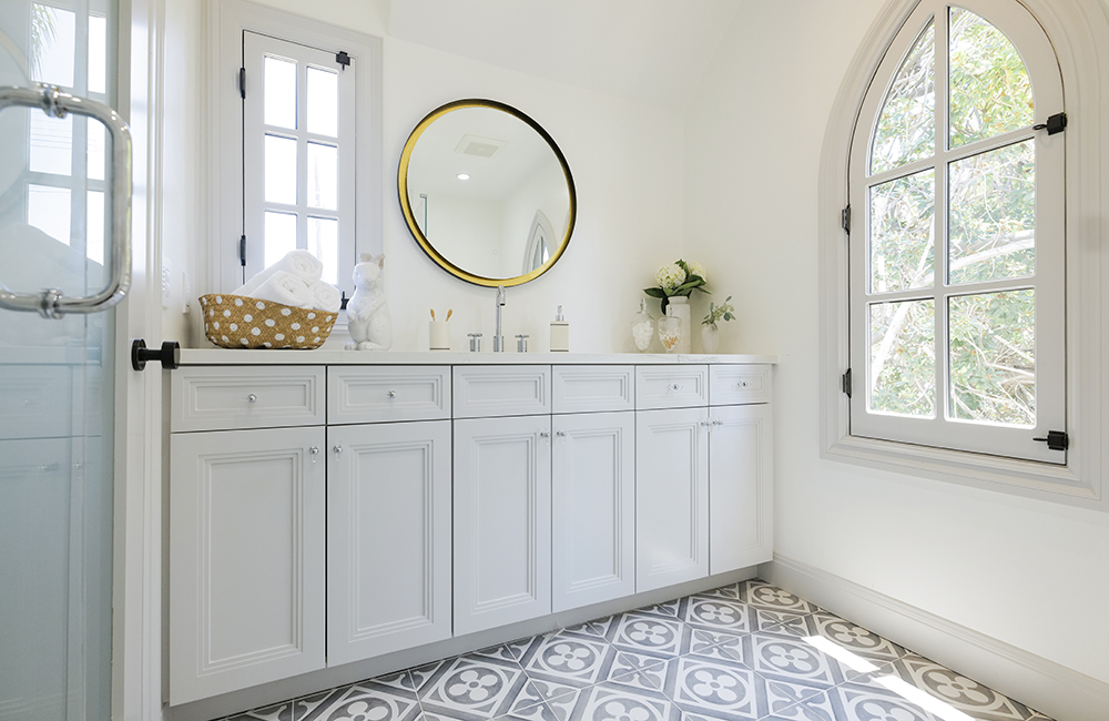 White bathroom with lots of light, tiled floor and gold-rimmed mirror