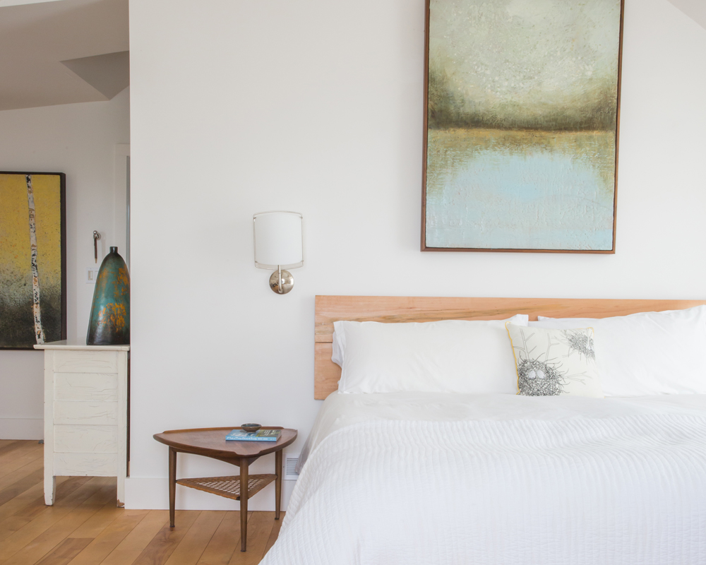 white bed in white bedroom with abstract painting above, white sconce, triangular wooden side table, white dresser around corner