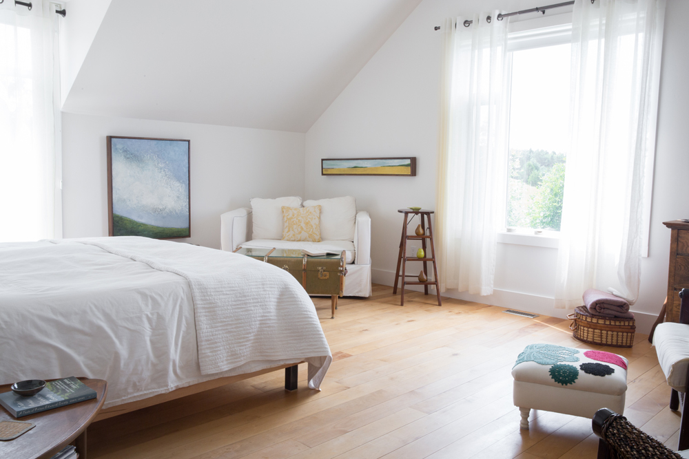 white bedroom with wood floor looking to window, ladder stool, sofa in corner, two pieces of art