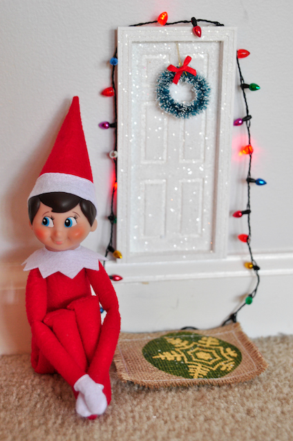 Elf on the Shelf sitting next to mini door with Christmas lights