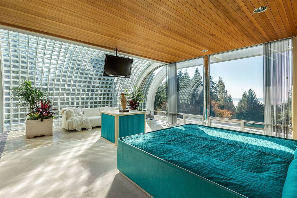 Bright, airy master bedroom with blue sheets and floor-to-ceiling windows overlooking forested area