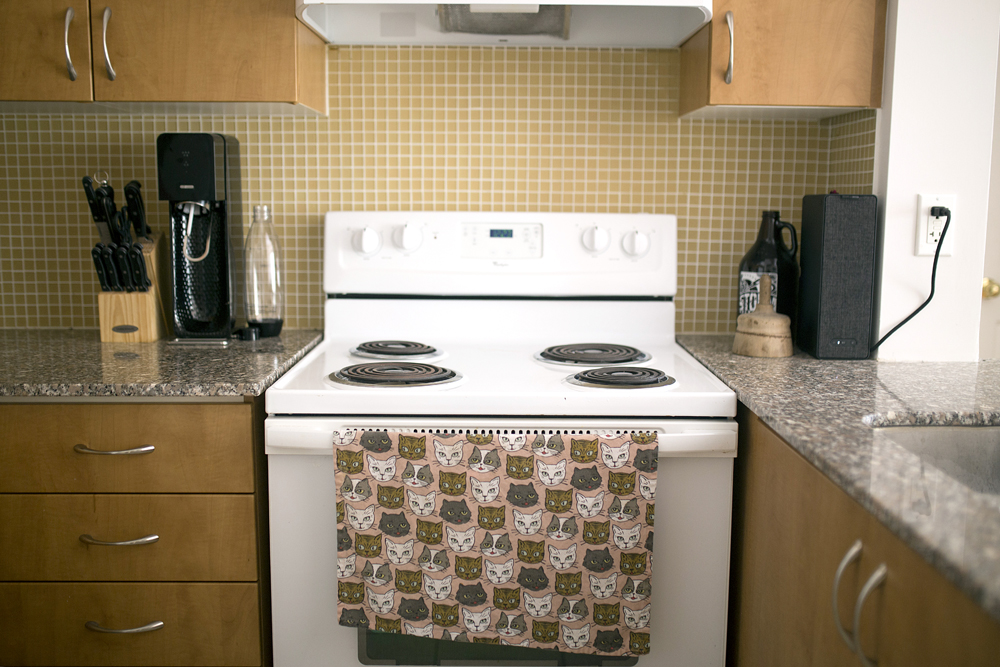 A kitchen stove closeup with various appliances on the countertop and a cat-patterned dish towel