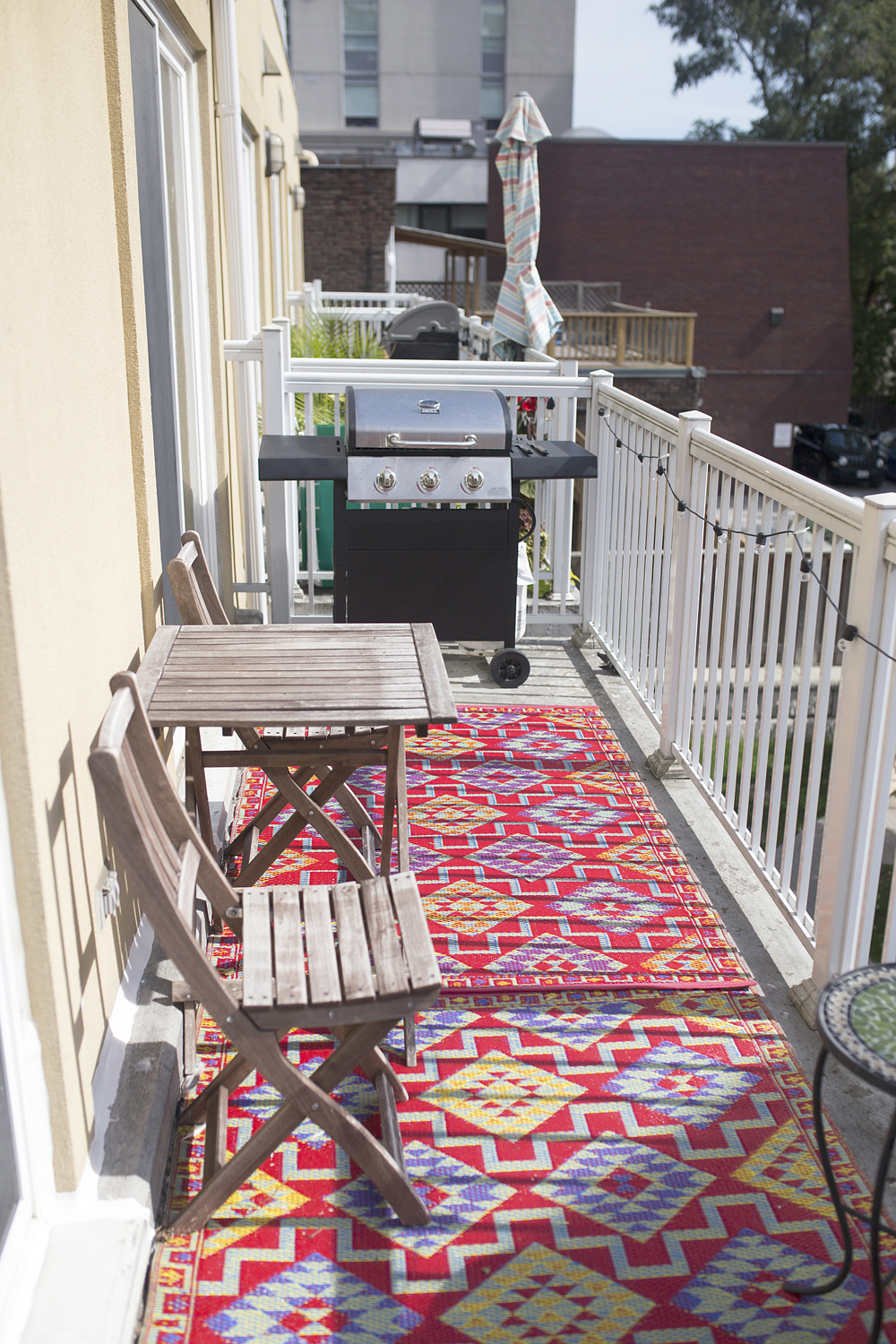 A narrow apartment balcony with an outdoor area rug, BBQ and patio furniture