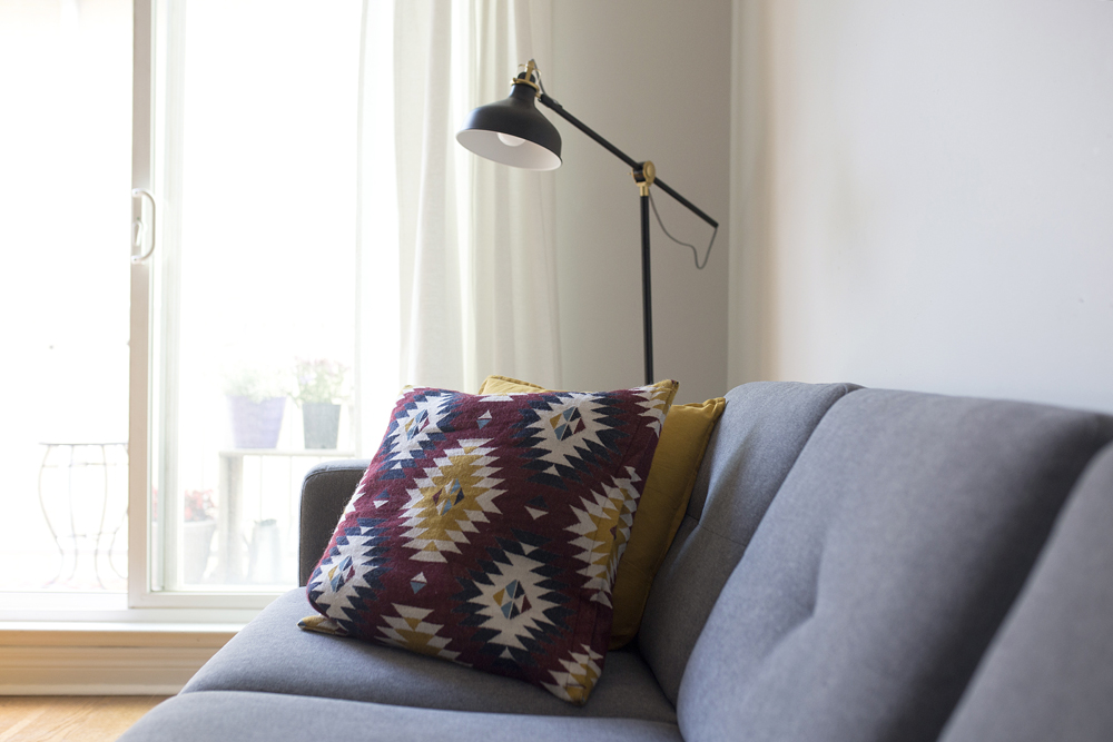 A close-up of a grey mid-century couch with patterned throw pillows and a narrow black lamp hanging over the couch arm
