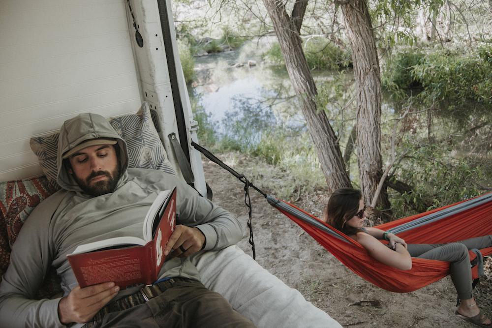 man reads in camper van while woman lays in attached hammock