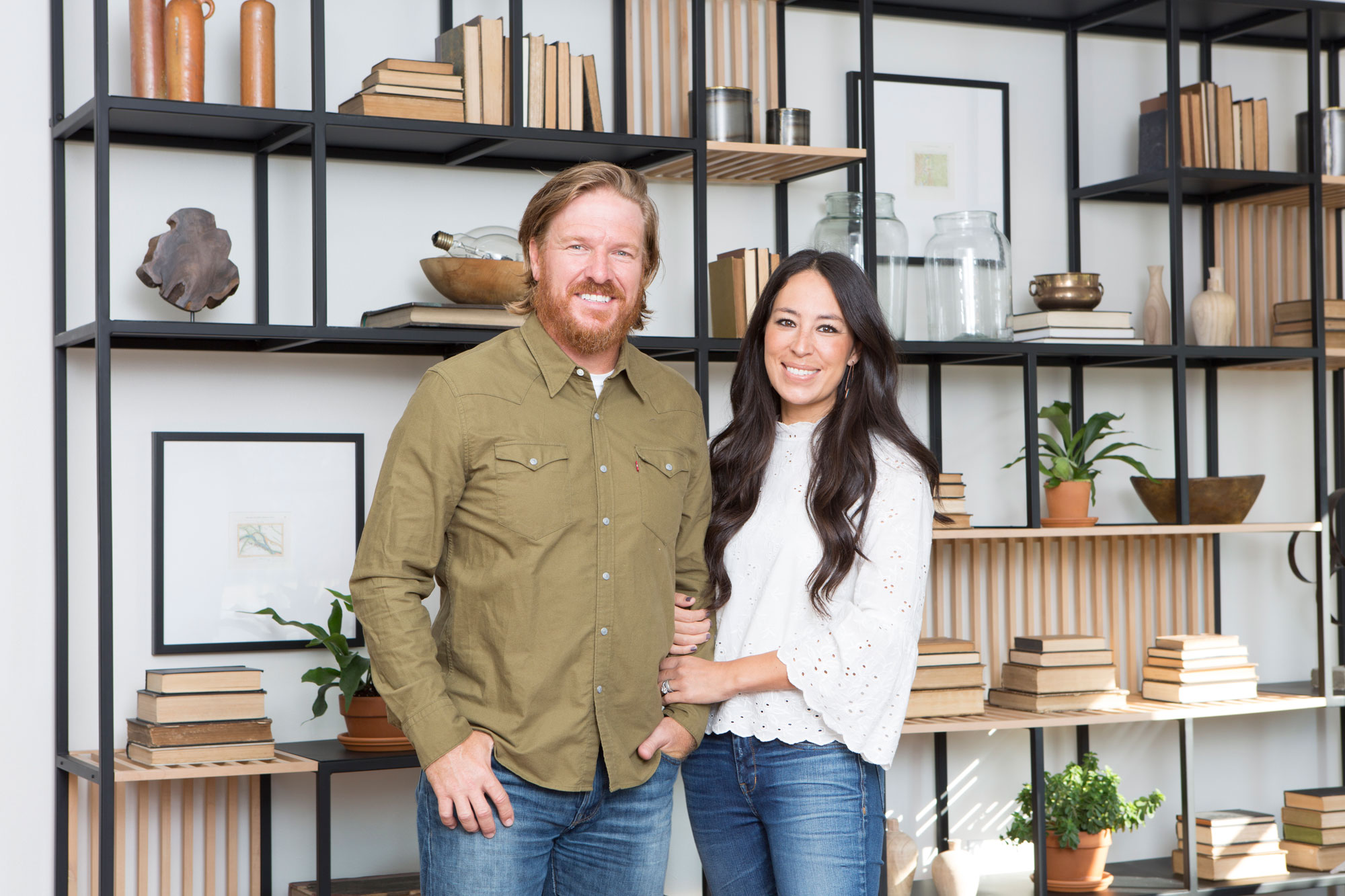 Chip and Joanna Gaines standing in front of a book shelf, posing for a photo.