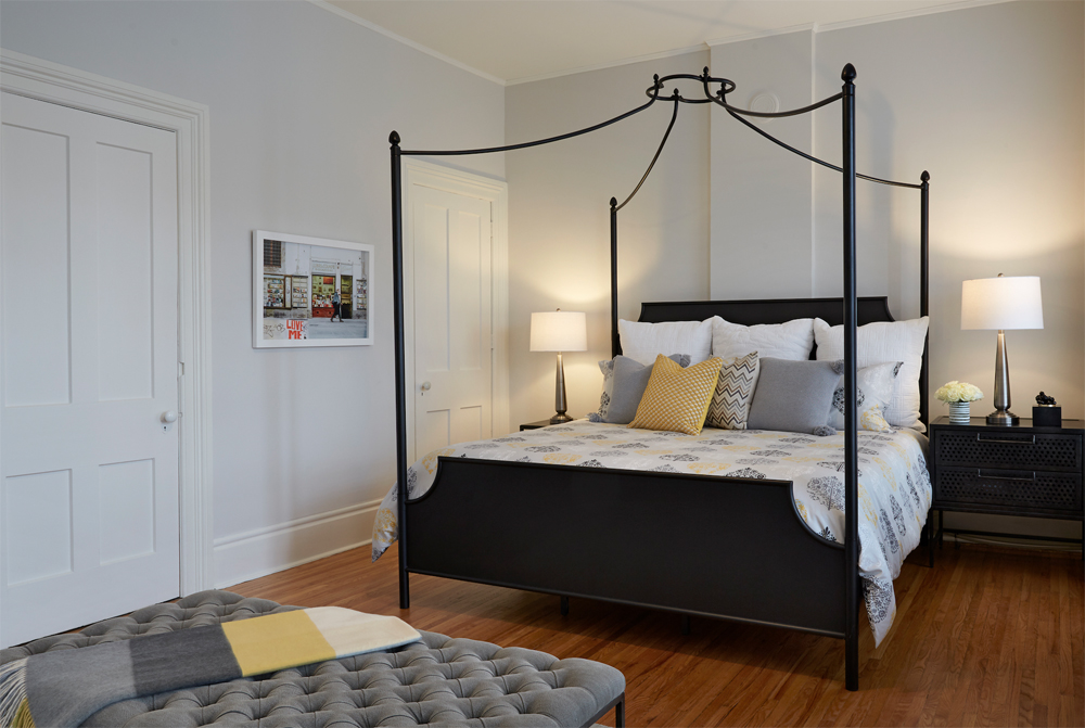 Large four-poster bed with grey and yellow accents, and hardwood floors throughout bedroom.