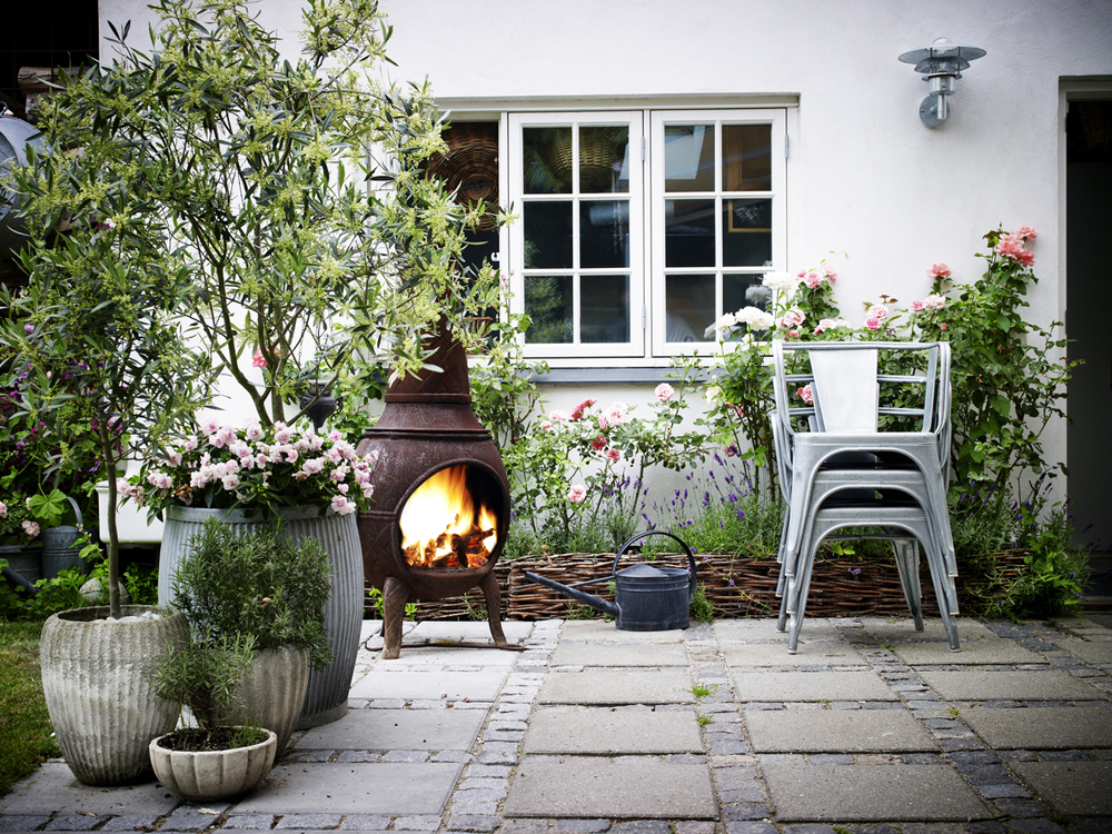 A fire ablaze in a chimenea in a backyard nook, with stone tiles on the floor and beautiful planted pots surrounding the space.
