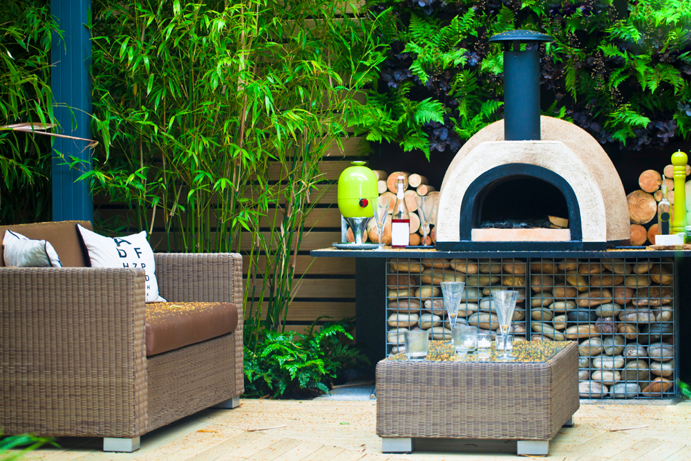 A backyard pizza oven sitting on-top of a beautiful stone table, with modern patio furniture and greenery in the background.