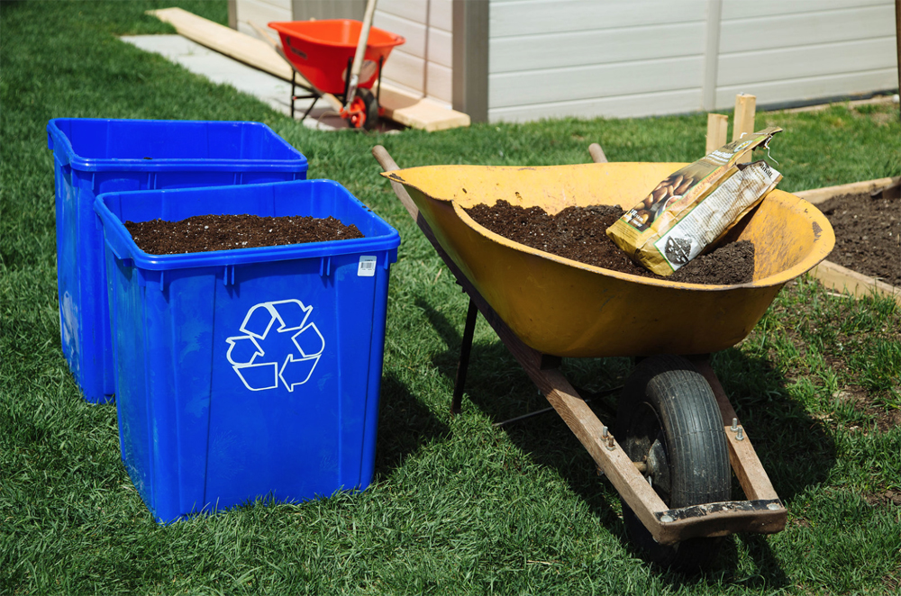 Two big blue recycling boxes filled with gardening soil and standing beside a large wheel barrel, also filled with gardening soil and a pack of potatoes.
