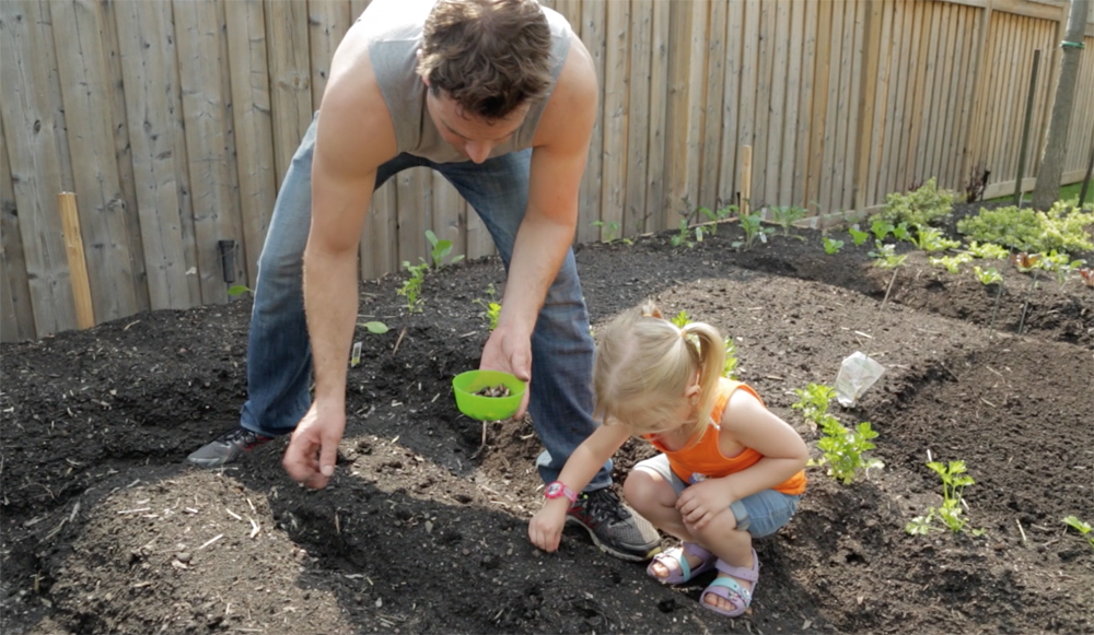Scott McGillivray planting in a backyard garden with a little girl helping him put seeds in the ground.