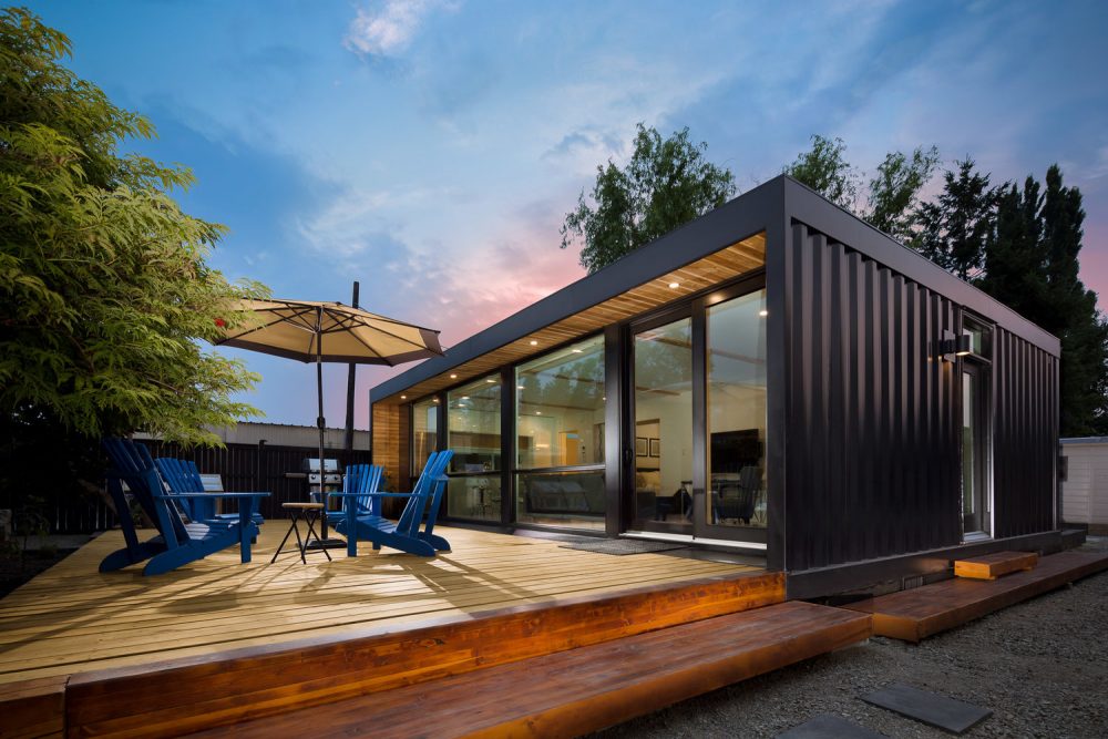 Black container home with large wooden deck featuring blue Muskoka chairs