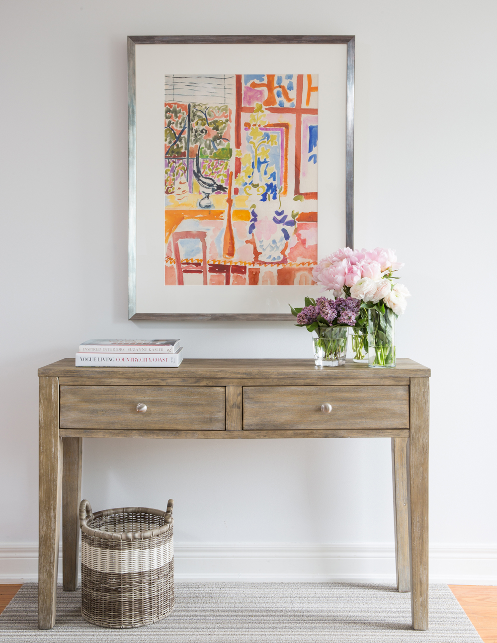 two drawer wood table, framed painting, basket below, Vogue Living bottom book on table