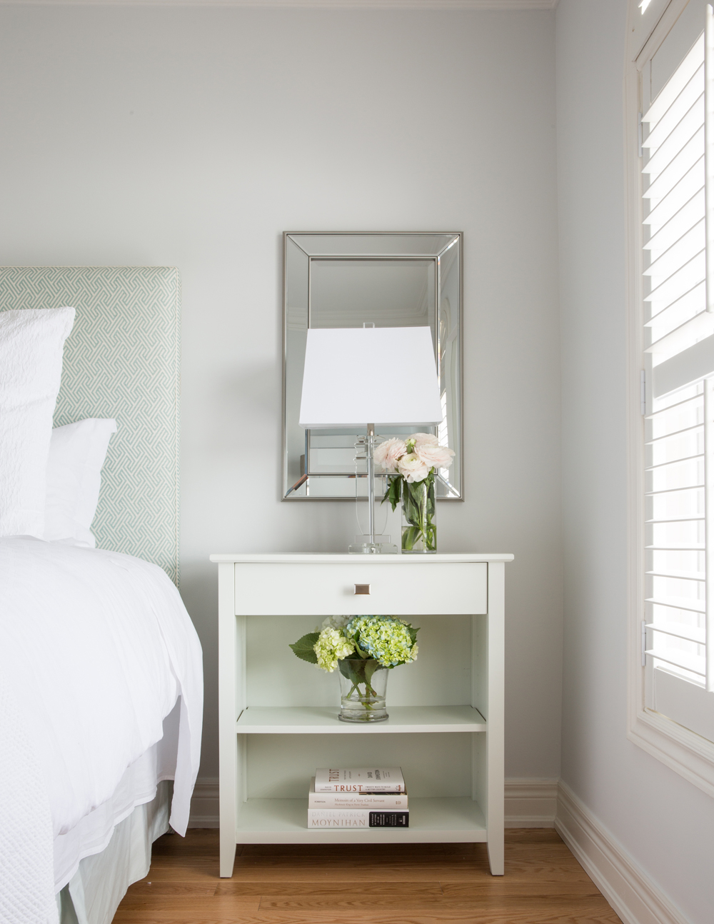 white bedside table with square drawer pull, two floral arrangements, three books on lower shelf