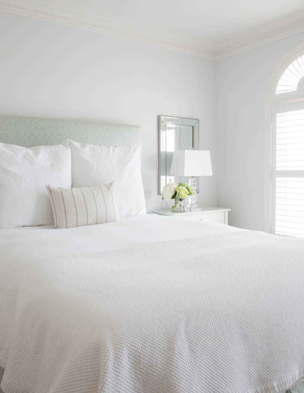 white bed with three cushions, light green patterned headboard, mirror behind lamp on white sidetable with flowers, shutters