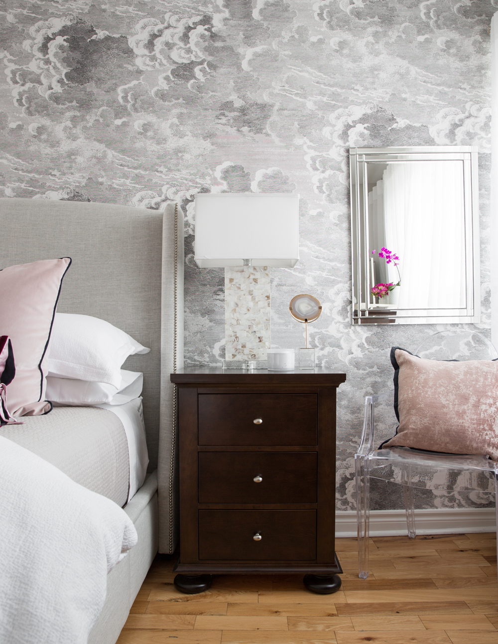 dark bedside table in front of cloud wallpaper beside clear chair with pink cushion
