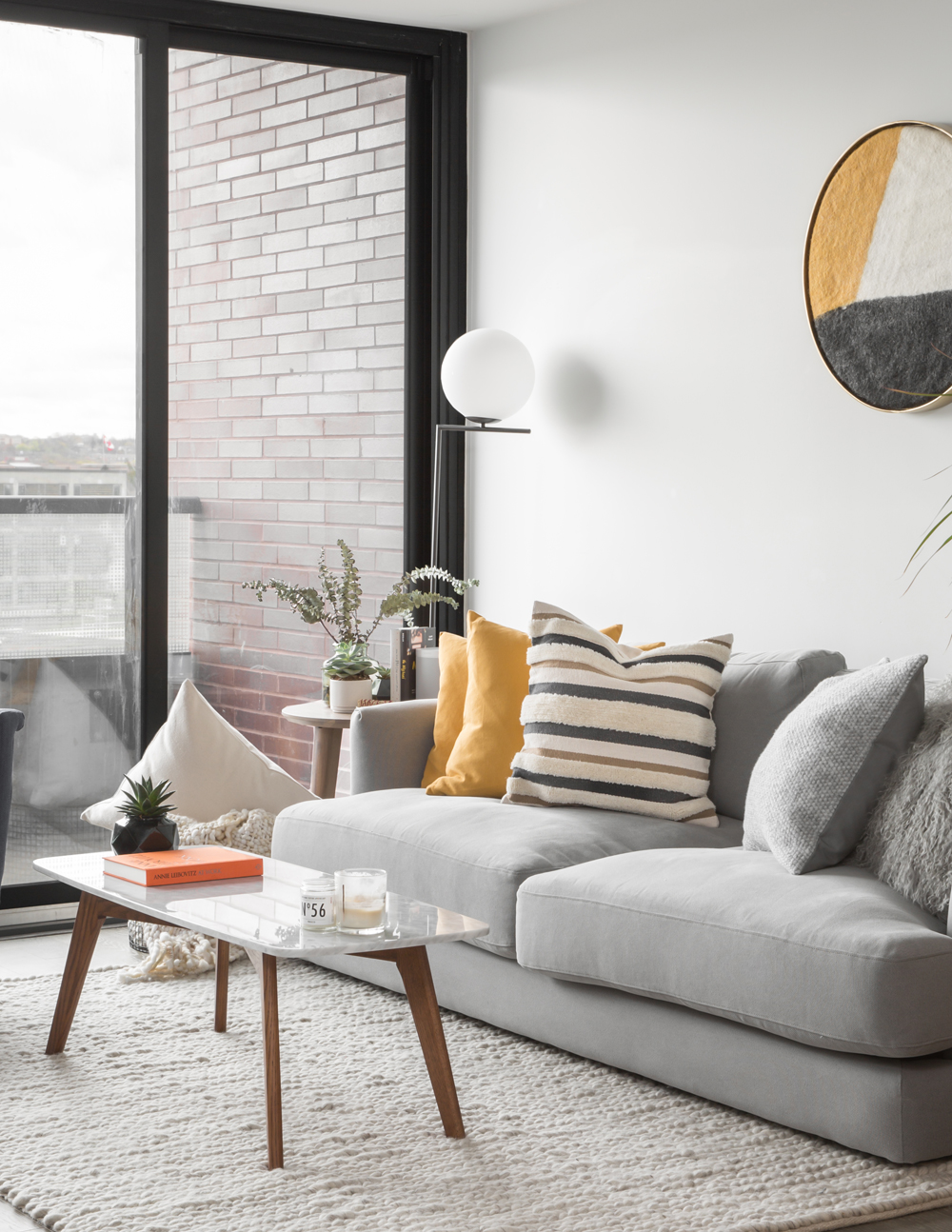 corner of condo with grey sofa, two mustard cushions on it, round bulb floor lamp in corner