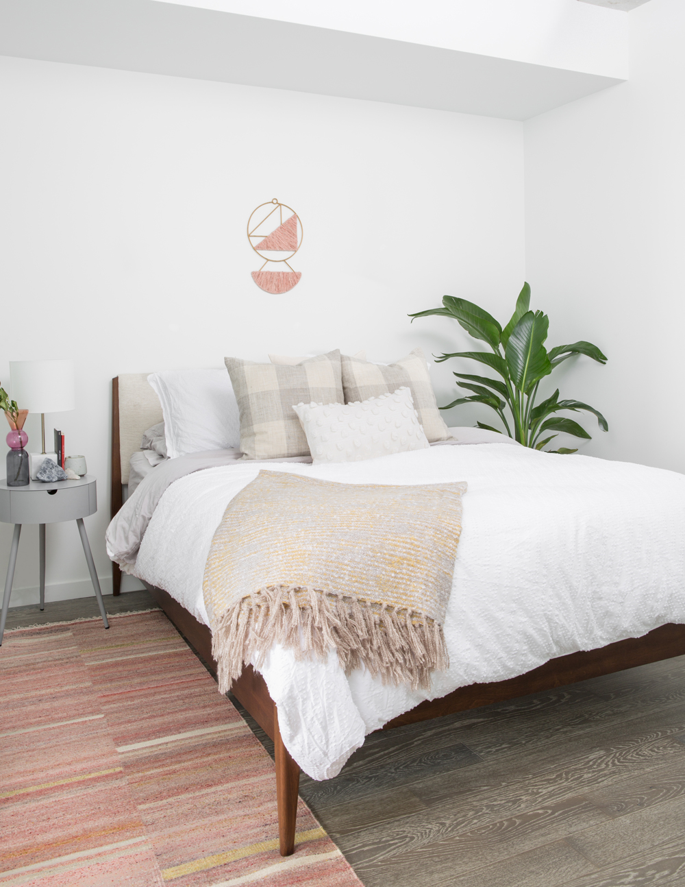 white bedroom with green plant in back right corner and small pink wall hanging