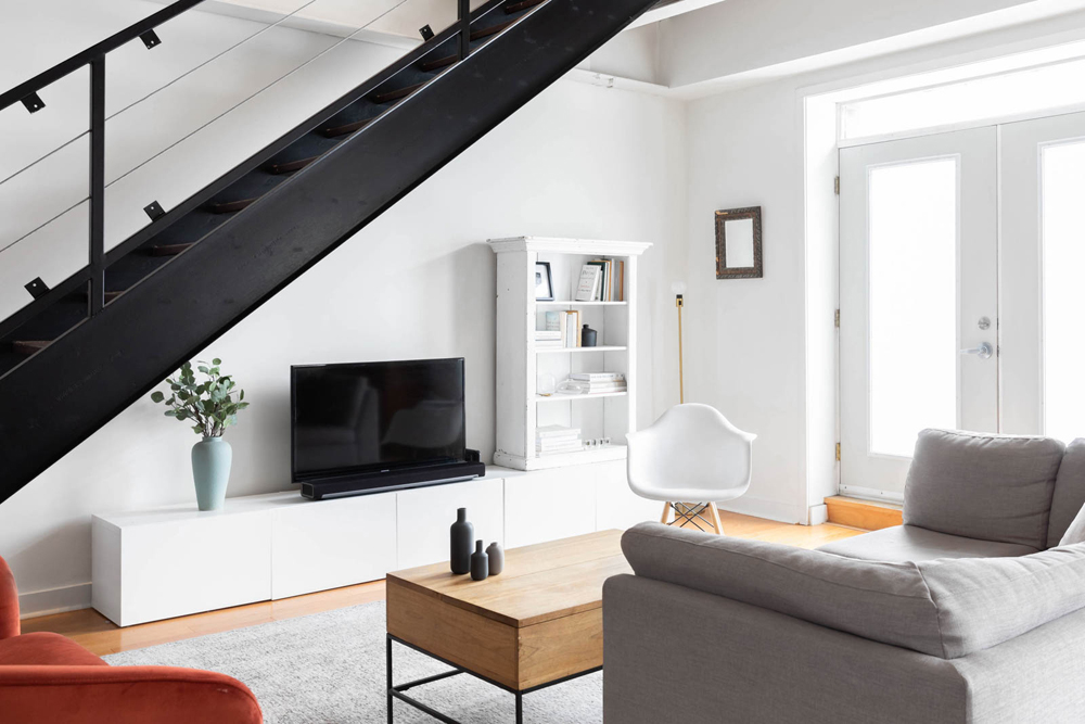 A TV under the black industrial-style staircase in the living room with white and grey furniture