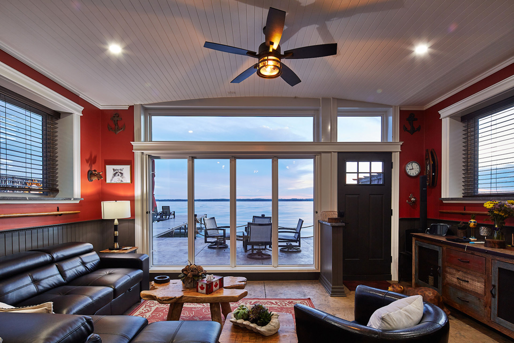 Cozy boathouse living room with dark leather sectional couch and armchair, a live edge wood coffee table, wooden credenza, red walls, white wood panel ceiling, dark metal industrial face, a red woven rug, and Magic™ windows and Magic Window Wall™ looking out to a patio set overlooking a lake at sunset