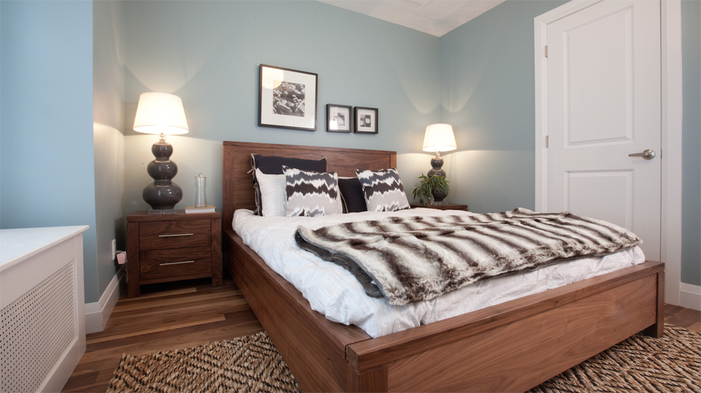 Renovated bedroom with pale blue walls, wood bed frame and hardwood flooring with patterned rug