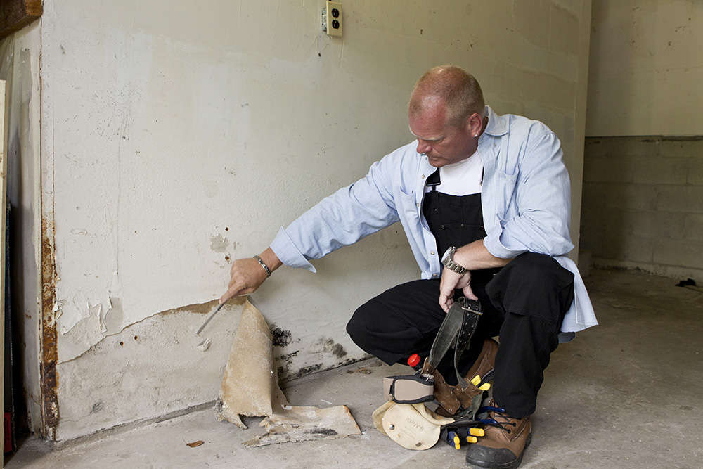 Mike Holmes in a basement peeling away wallpaper to reveal mould growth