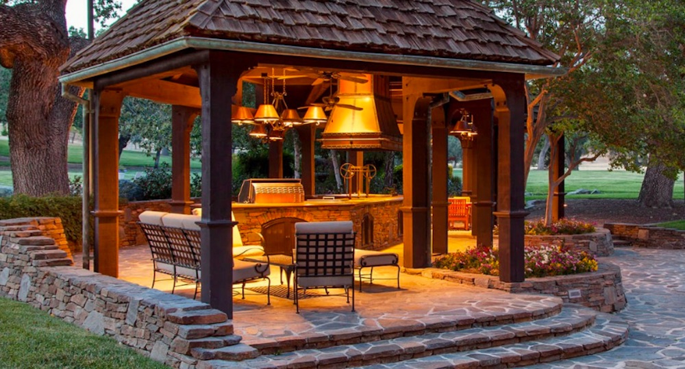 A brick and wood gazebo housing patio furniture and a large stone barbecue pit